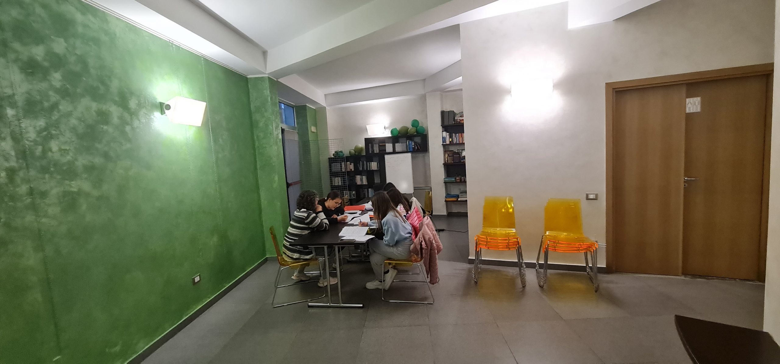 Sala con tavolo dove sono sedute quattro bambine e una donna, sedie arancioni e una porta in legno sulla destra, un muro verde a sinistra, in fondo una libreria.