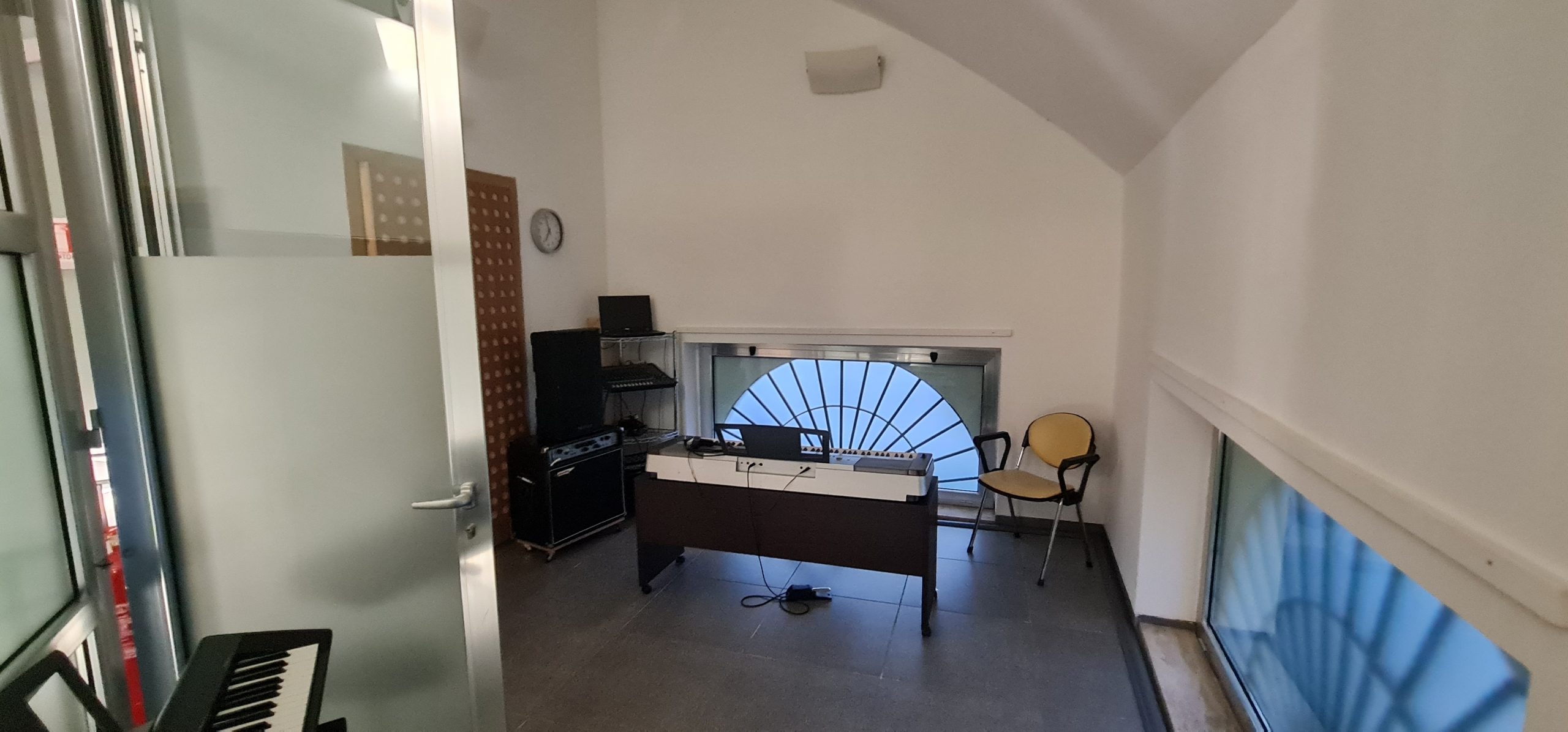 Sala con a sinistra una porta aperta, al centro una tastiera e ai lati casse audio e due grandi finestre semicircolari che si trovano in basso sui muri laterali bianchi.