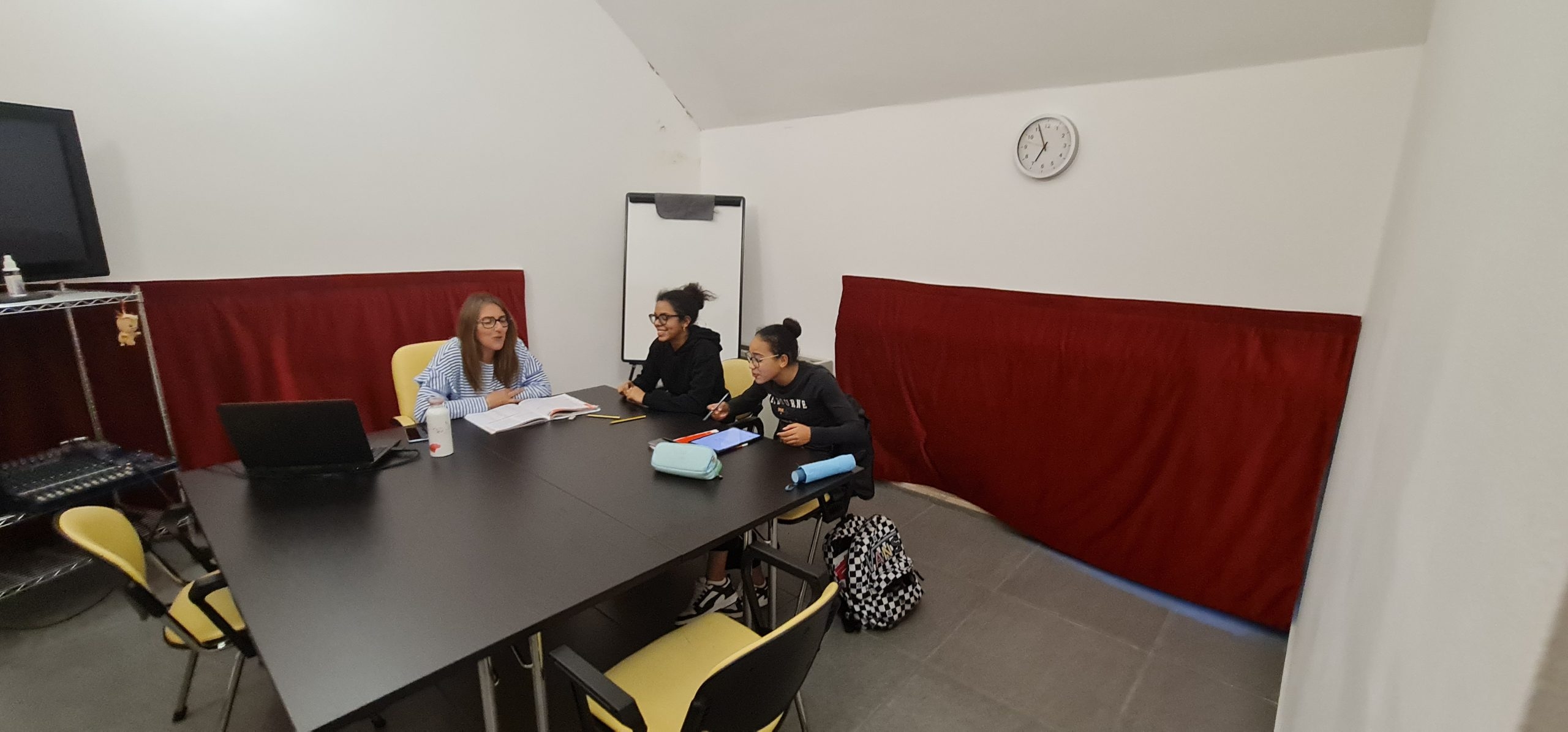 Tre ragazze studiano sedute a un tavolo nero con sedie gialle e su cui ci sono alcuni fogli. Il tavolo è al centro di una sala con le pareti bianche e delle lunghe tende rosse in basso, un orologio bianco sul muro di destra, una lavagna a fogli mobili in fondo al centro e una tv nera su un mobile sulla sinistra.