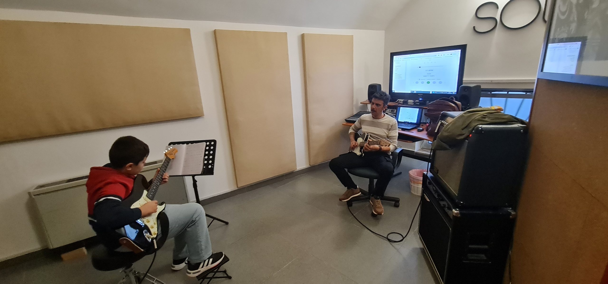 Aula insonorizzata dove un bambino su una sedia a sinistra e un uomo su una sedia a destra stanno suonando due chitarre, alle spalle dell'uomo si trova un monitor acceso.