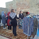 Foto dell'evento "CLOTHES SWAP: BARATTIAMO I NOSTRI VESTITI" Un gruppo di persone, principalmente donne, è riunito intorno a un appendiabiti con vari capi di abbigliamento colorati. Alcune persone stanno esaminando i vestiti. L'area è all'aperto, con un tappeto decorato sotto i piedi e un edificio in pietra sullo sfondo. L'atmosfera è conviviale e rilassata.