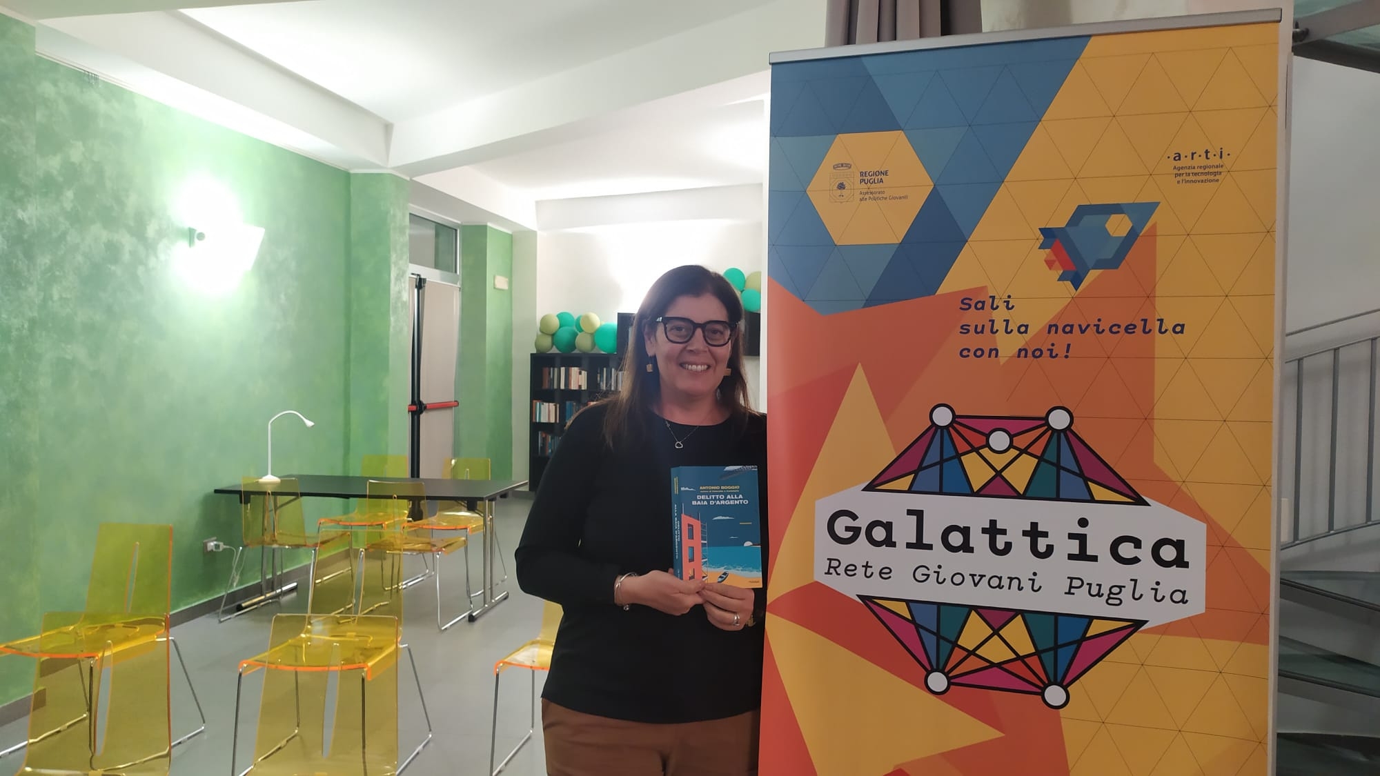 A destra il roll up del nodo di Galattica di Mesagne, accanto una donna in piedi mostra la copertina di un libro e a sinistra sedie gialle di plastica trasparente in una sala con pareti verdi e soffitto bianco.