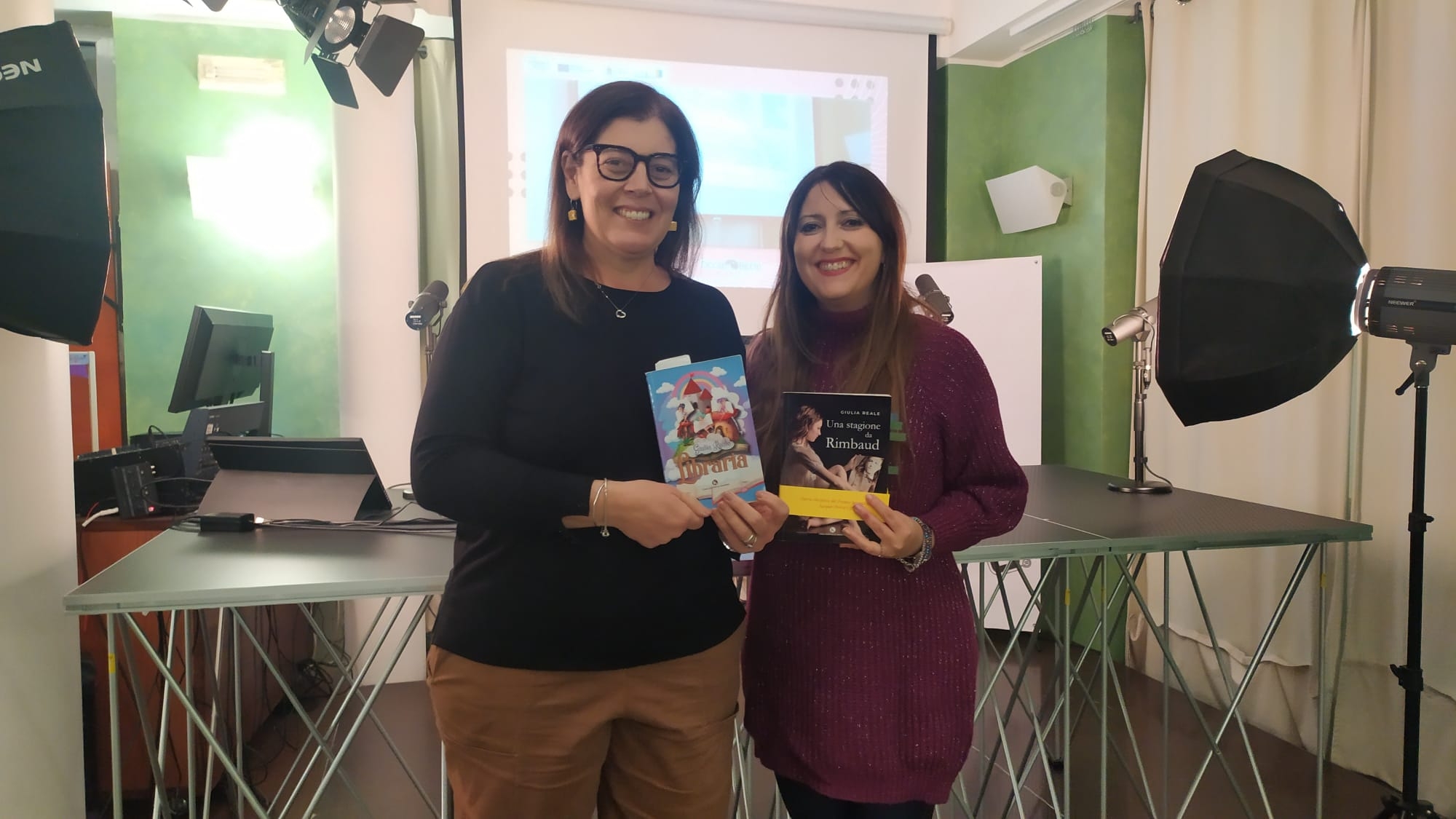 Due donne in piedi una accanto all'altra ognuna con un libro tra le mani mostrano le copertine, dietro di loro un tavolo grigio e una lavagna bianca in una sala con tende bianche e pareti verdi.