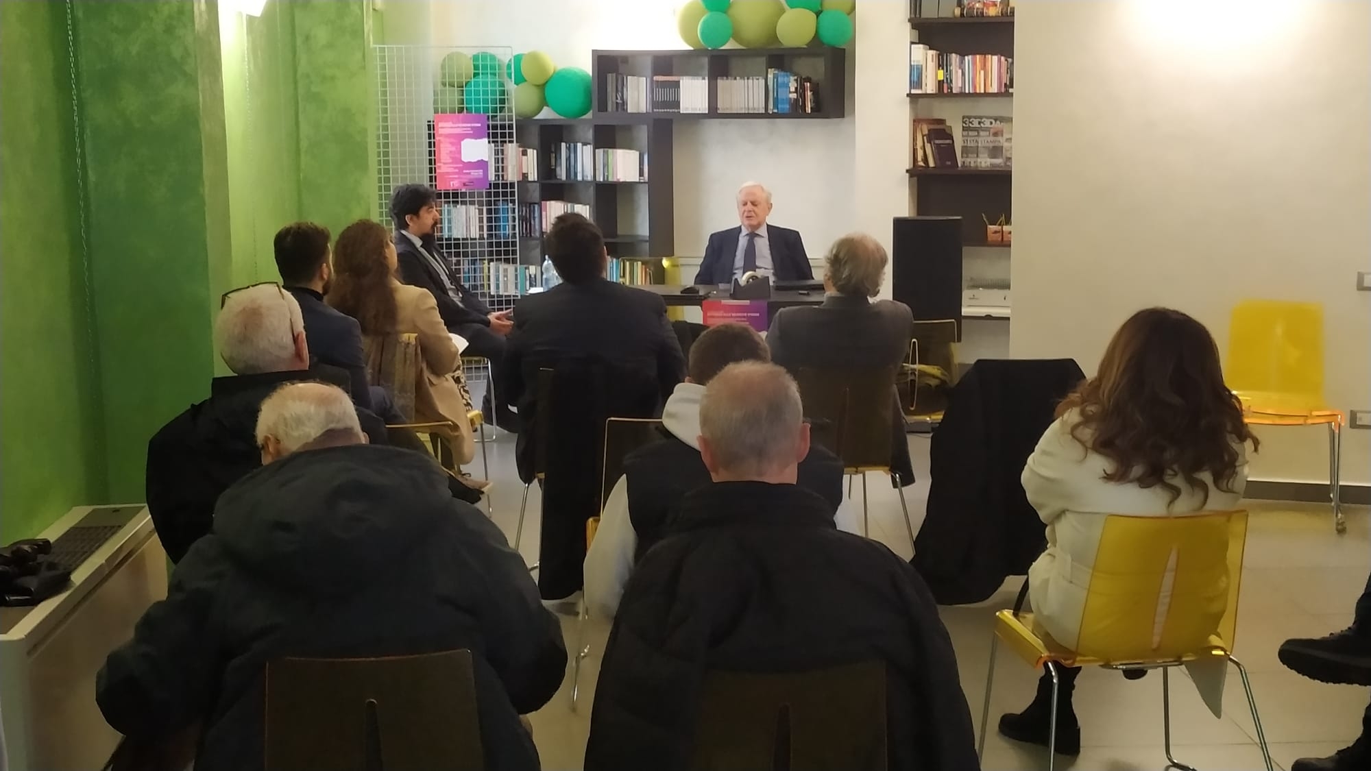 Persone di spalle sedute su siede gialle, in fondo un uomo dietro a una scrivania di fronte a loro e dietro uno scaffale addossato alla parete verde a sinistra e bianca a destra.