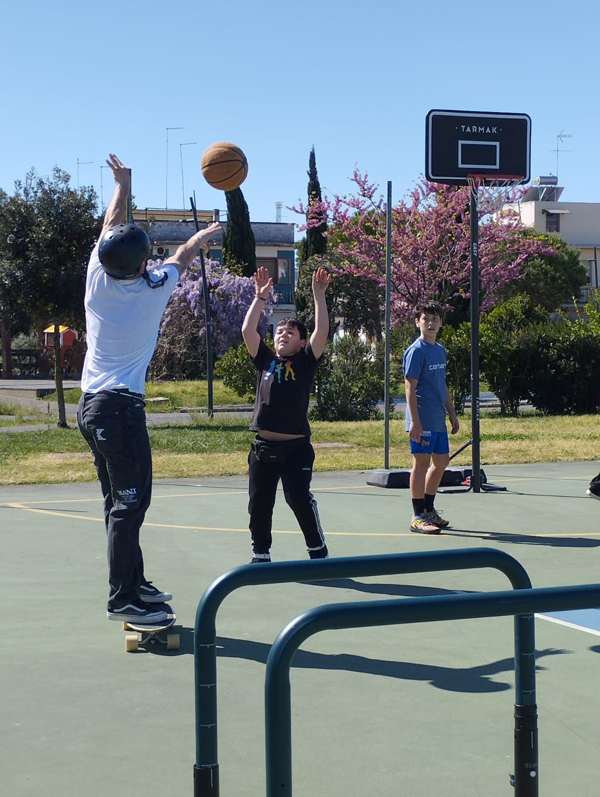 Ragazzo su uno skateboard di spalle si trova su un campo da basket e lancia una palla verso il canestro, dietro due ragazzi lo guardano e il primo è con le braccia alzate, In fondo ci sono alberi fioriti e il cielo azzurro.