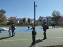 Ragazzi e bambini su un campetto con una parte colorata di blu e una rampa, in fondo ci sono alberi fioriti e il cielo azzurro.