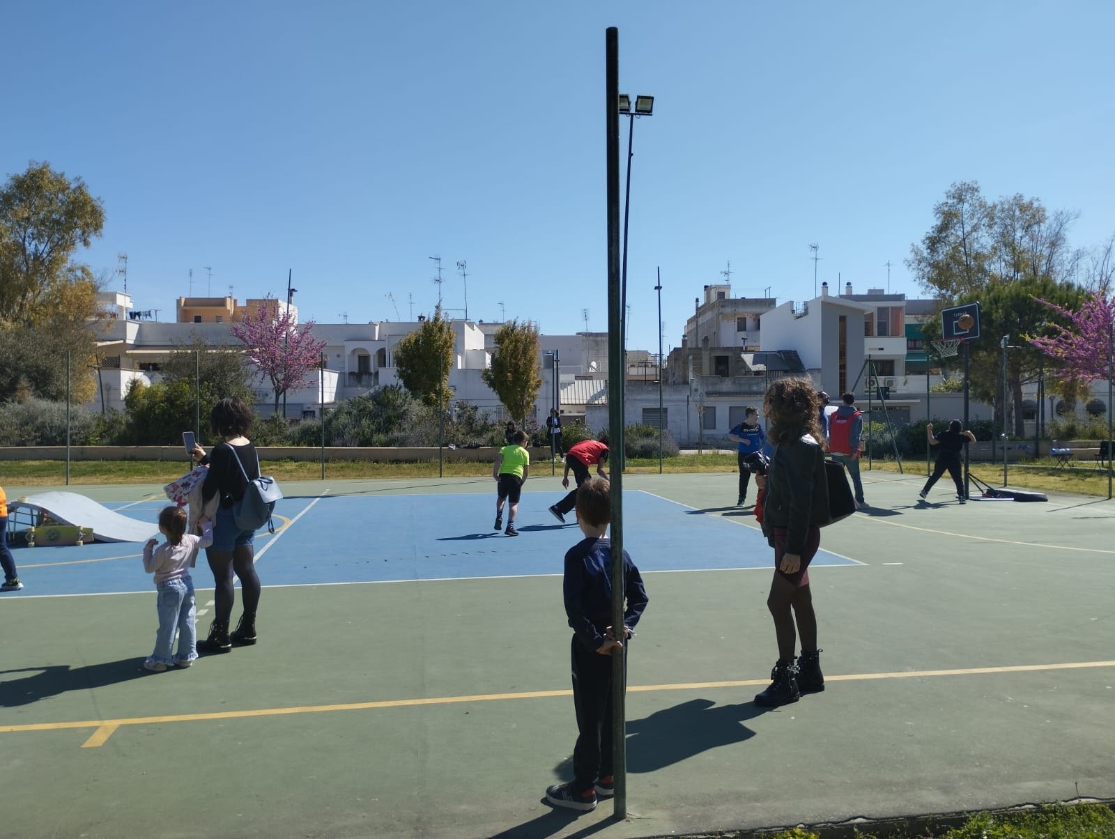 Ragazzi e bambini su un campetto con una parte colorata di blu e una rampa, in fondo ci sono alberi fioriti e il cielo azzurro.