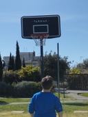 Ragazzo visto di spalle si trova di fronte a un canestro, in fondo ci sono siepi e alberi verdi e il cielo azzurro.