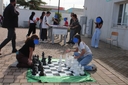 in primo piano una scacchiera gigante in terra con scacchi grandi e due bambini ai lati della scacchiera che stanno giocando. In secondo piano, biliardini con ragazzi e ragazze ai lati che stanno giocando. ai lati dell'immagini ci sono, sul lato sinistro un signore con gli occhiali che sta cmminando verso il biliardino. Dal'altro lato una ragazza che guarda il suo cellulare.