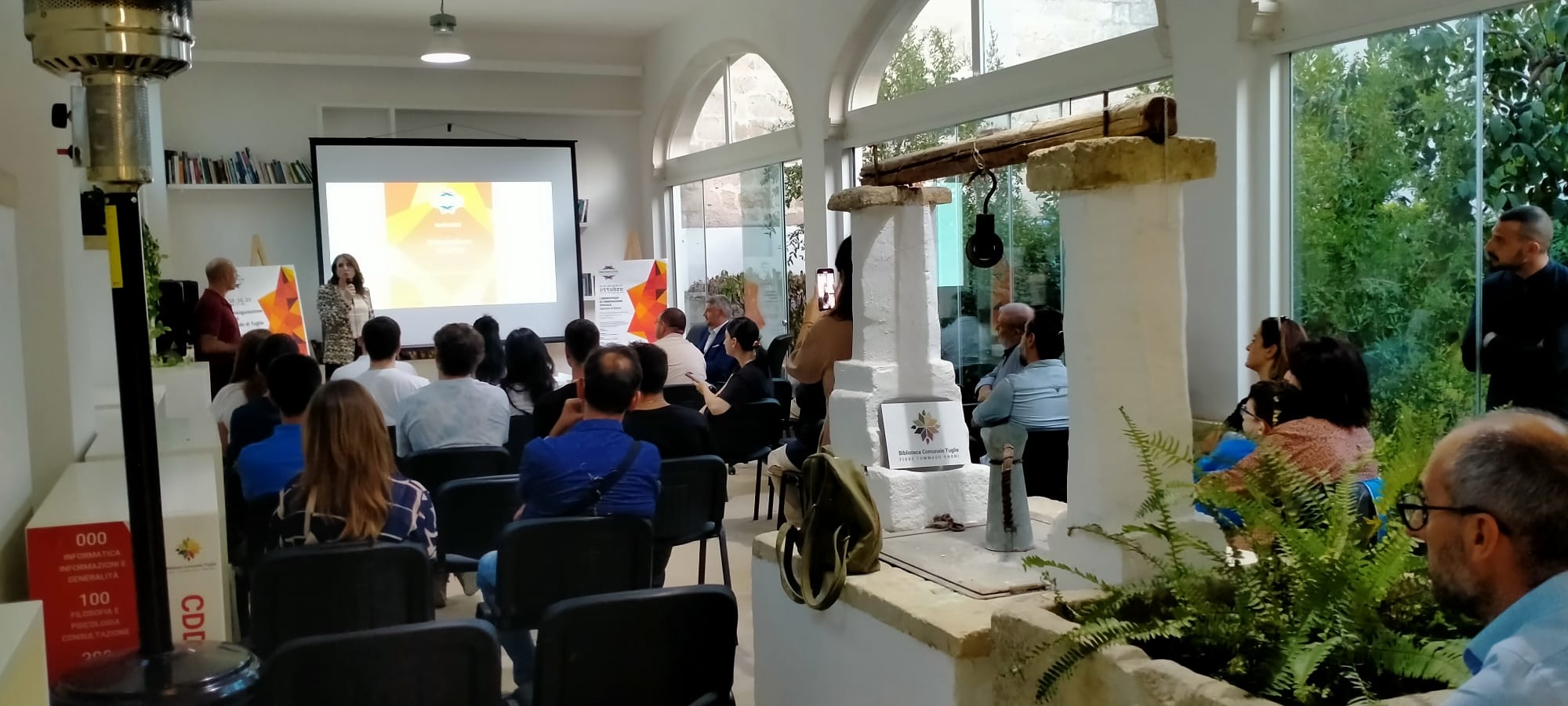 Persone sedute su sedie in una stanza davanti a una presentazione sul proiettore, con una donna che presenta