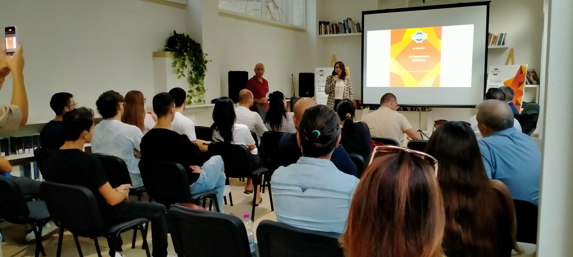 Persone sedute su sedie in una stanza davanti a una presentazione sul proiettore, con una donna che presenta