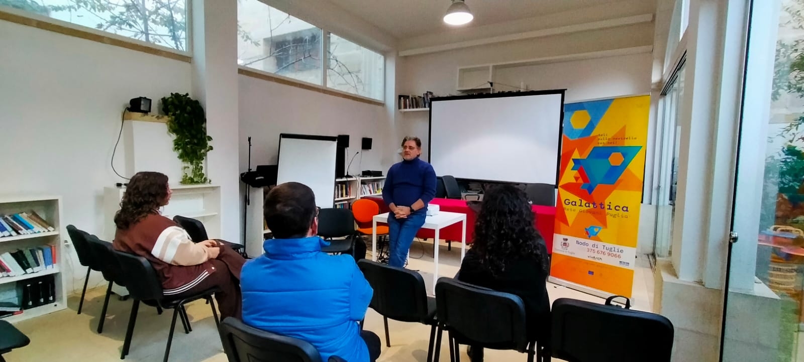 Persone sedute su sedie in una stanza davanti a una presentazione sul proiettore, con un docente che presenta