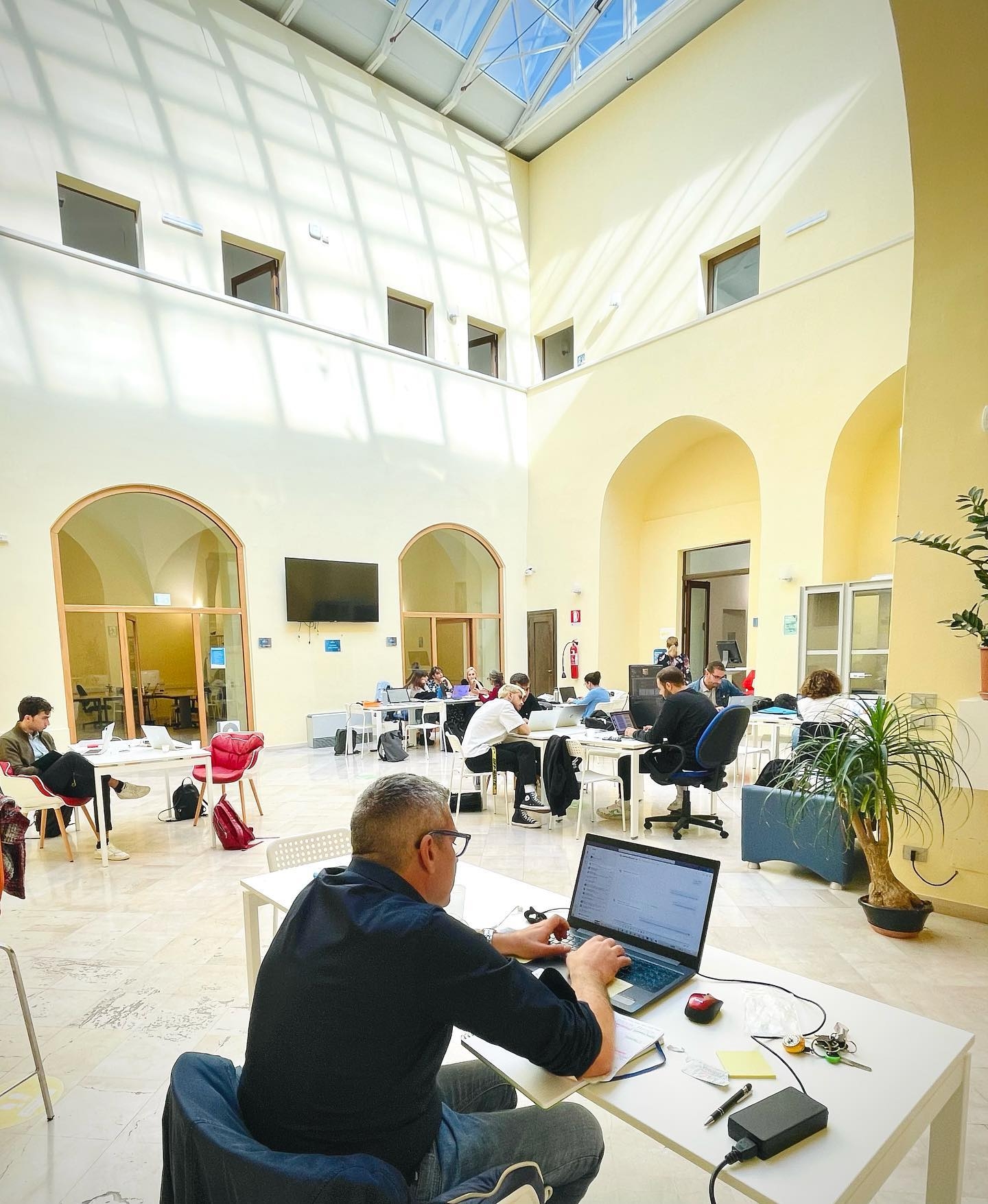 L'immagine mostra l'ampia e luminosa sala di coworking di Palazzo Guerrieri, sede del Nodo Galattica di Brindisi e le sue numerose postazioni dove alcuni utenti studiano e lavorano.