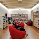 Sala lettura con pouf rossi su cui ci sono ragazze e ragazzi, scaffali pieni di libri sulle pareti.