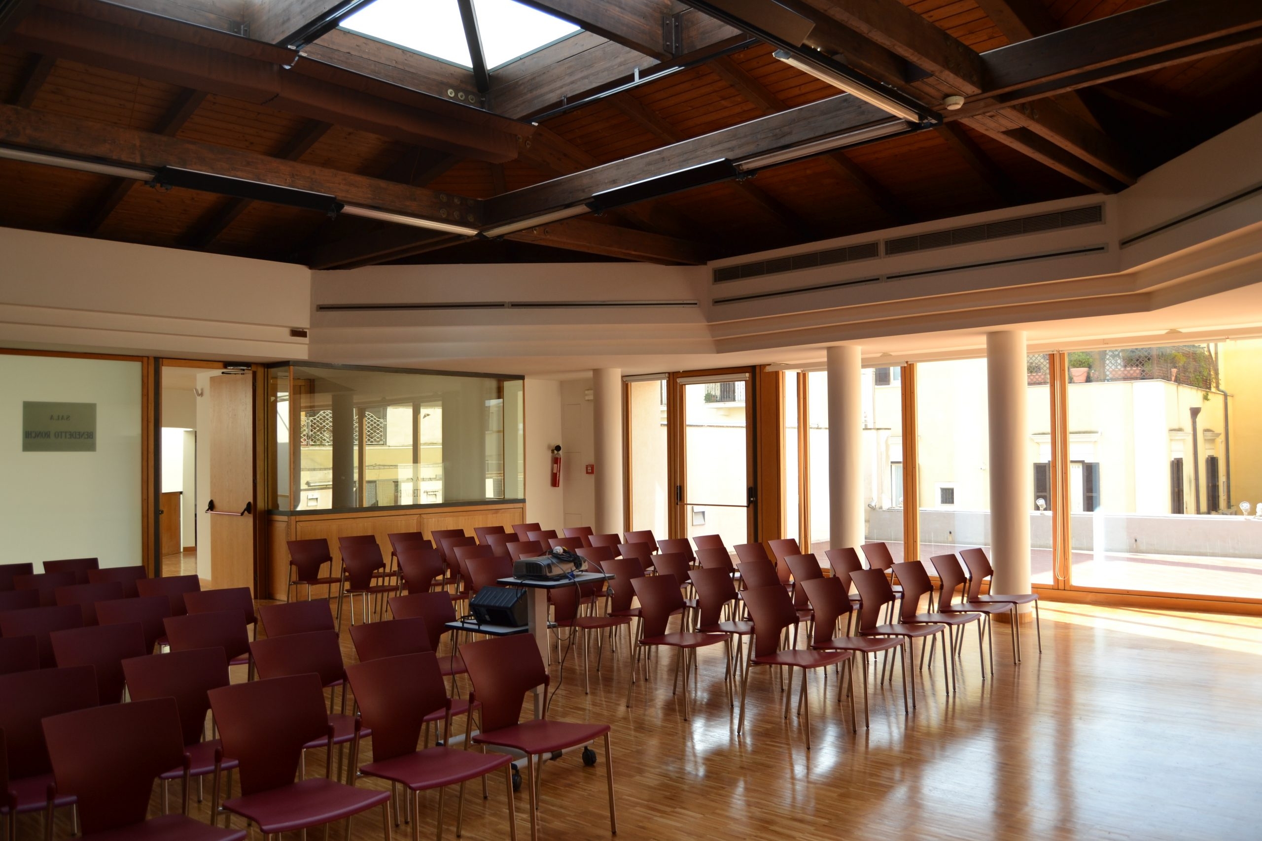 Sala con sedie rosse disposte in fila, tettoia in legno con apertura centrale e vetrate laterali