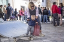 Un infante sorretto da una donna sta in piedi su uno skateboard, dietro ci sono una rampa e tante persone di spalle.