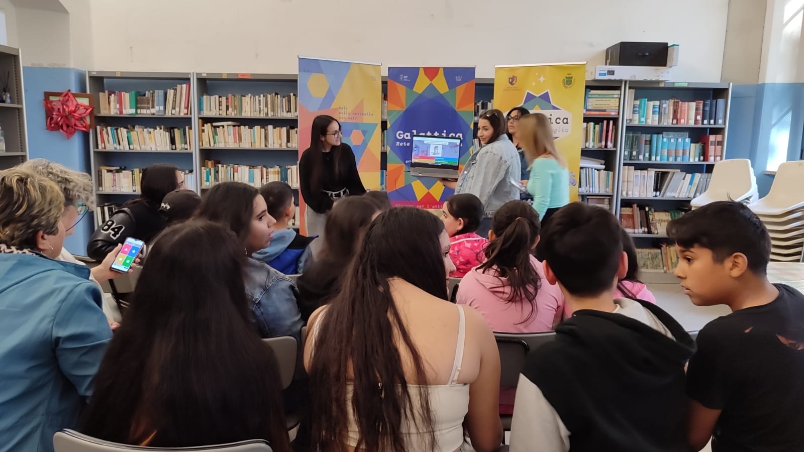 Ragazzi di spalle in primo piano, al centro quattro ragazze alzate davanti roll-up di Galattica