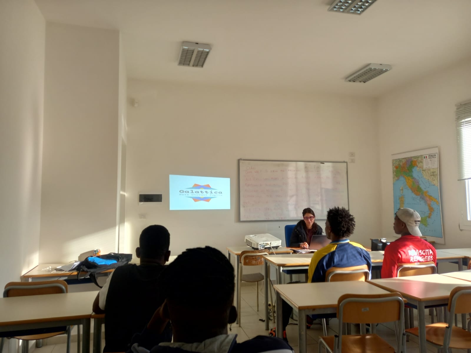 è una classe di una scuola co alcuni banchi. Ci sono quattro ragazzi seduti di spalle e alla cattedra è seduta una ragazza con gli occhiali che sta proiettando qualcosa sul muro dal suo computer. Sul muro logo di galattica. Dietro alla ragazza seduta alla cattedra, sull aparete, lavagna con delle scritte rosse sopra. Nella parete adiacente cartina geografica dell'Italia. Accanto alla cartina, di intravedono e veneziane di una finestra.