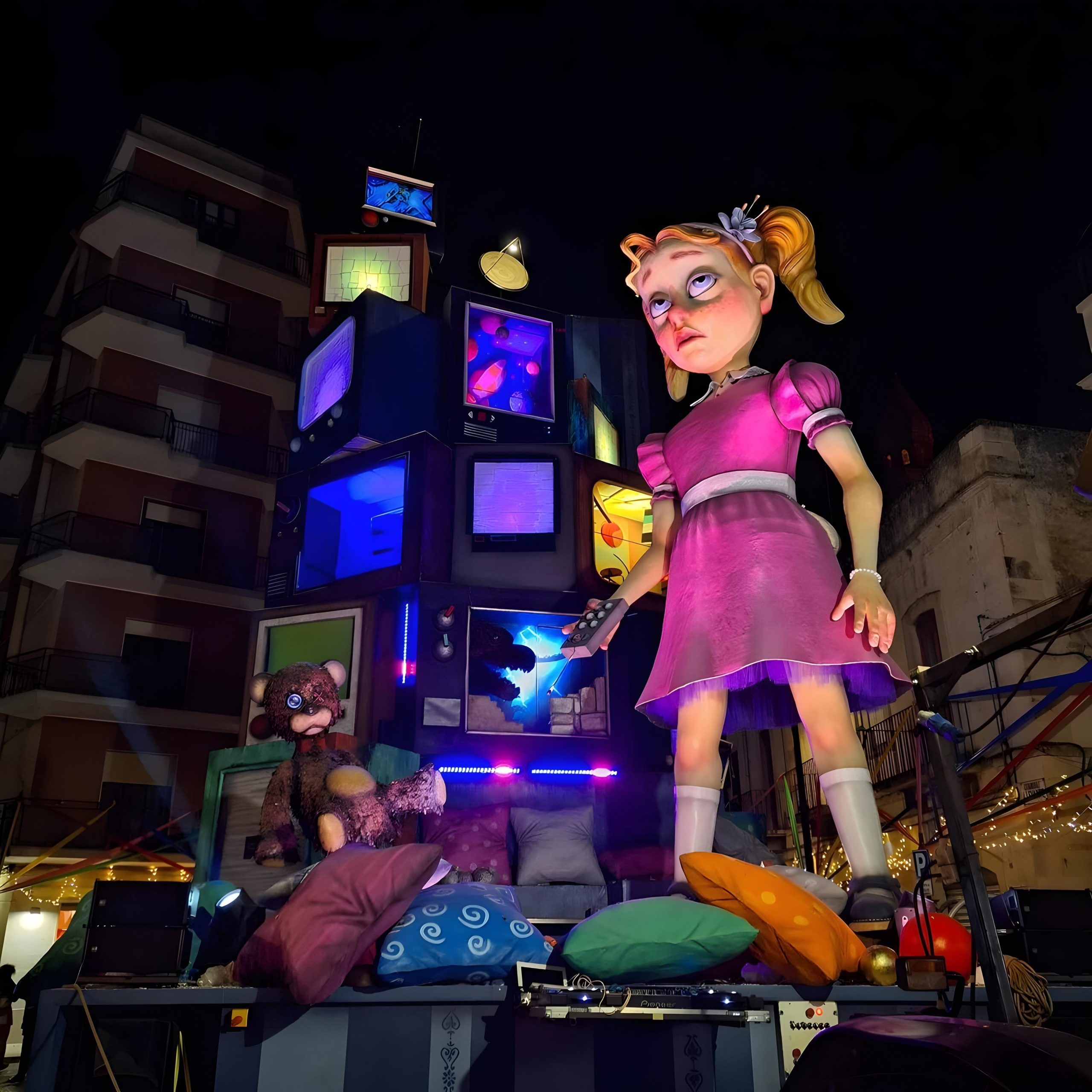 Maggie, scultura gigante di una bambina vestita di rosa, illuminata da luci colorate, circondata da televisori e un orsetto di peluche, durante il Carnevale di Putignano