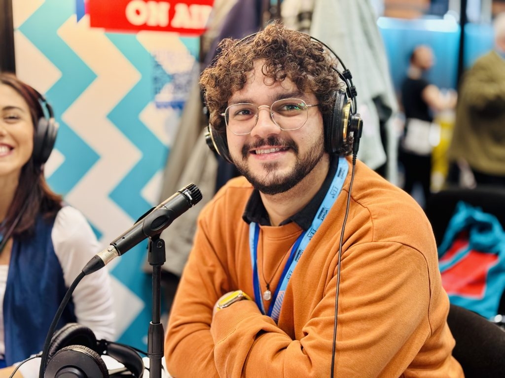 Primo piano di Alessandro Cassotta, speaker di Mao FM 19.77, durante il Womex 2024. Indossa le cuffie e parla al microfono in una postazione radiofonica allestita per l'evento.