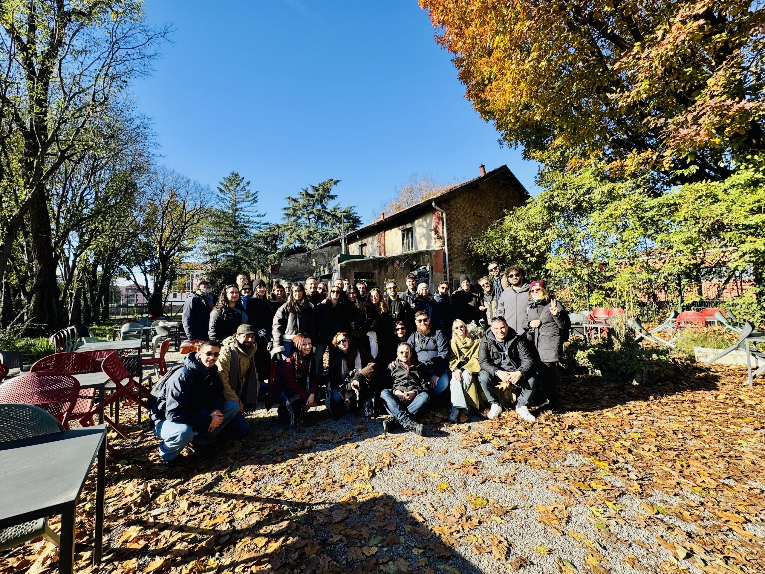 Foto di gruppo del team di Luoghi Comuni all'aperto a Milano, circondato da alberi autunnali e sedie rosse, con un edificio rustico sullo sfondo