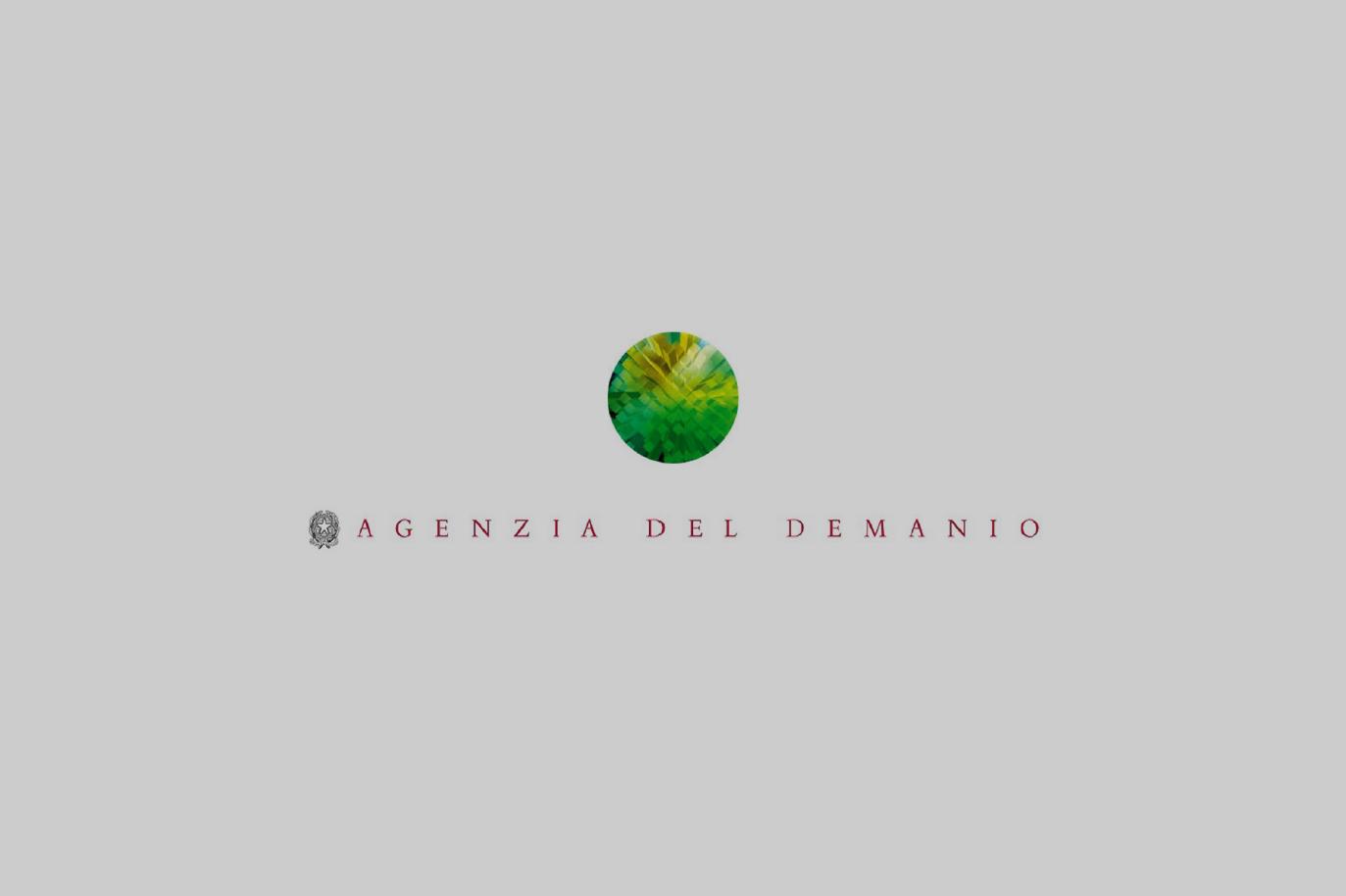 Logo ufficiale dell'Agenzia del Demanio su sfondo grigio. Al centro è presente un cerchio verde stilizzato con sfumature geometriche, che richiama elementi naturali e territoriali. Sotto il simbolo, il nome 'Agenzia del Demanio' è scritto in caratteri eleganti di colore rosso, con un piccolo stemma della Repubblica Italiana sulla sinistra. Questo logo rappresenta l'ente pubblico italiano responsabile della gestione e valorizzazione del patrimonio immobiliare dello Stato.
