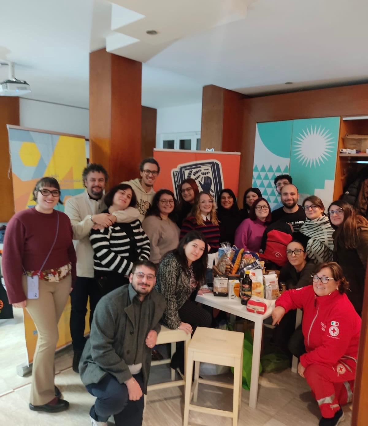 L’immagine mostra un gruppo affiatato di circa 15 persone (uomini e donne di diverse età) riunite in un ambiente interno luminoso. L'atmosfera è amichevole e informale; molti sorridono e sono in posa per una foto ricordo. Al centro della scena c'è un tavolo bianco carico di pacchi di cibo e generi alimentari.