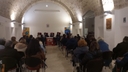 Presentazione del libro-platea di persone che ascoltano Platea di persone e tavolo centrale alla stanza. Dietro al tavolo sono sedute quattro persone e a fianco del tavolo, donna davanti a un leggio che legge qualcosa al microfono. Ai lati de tavolo ci sono due roll-up di Galattica.