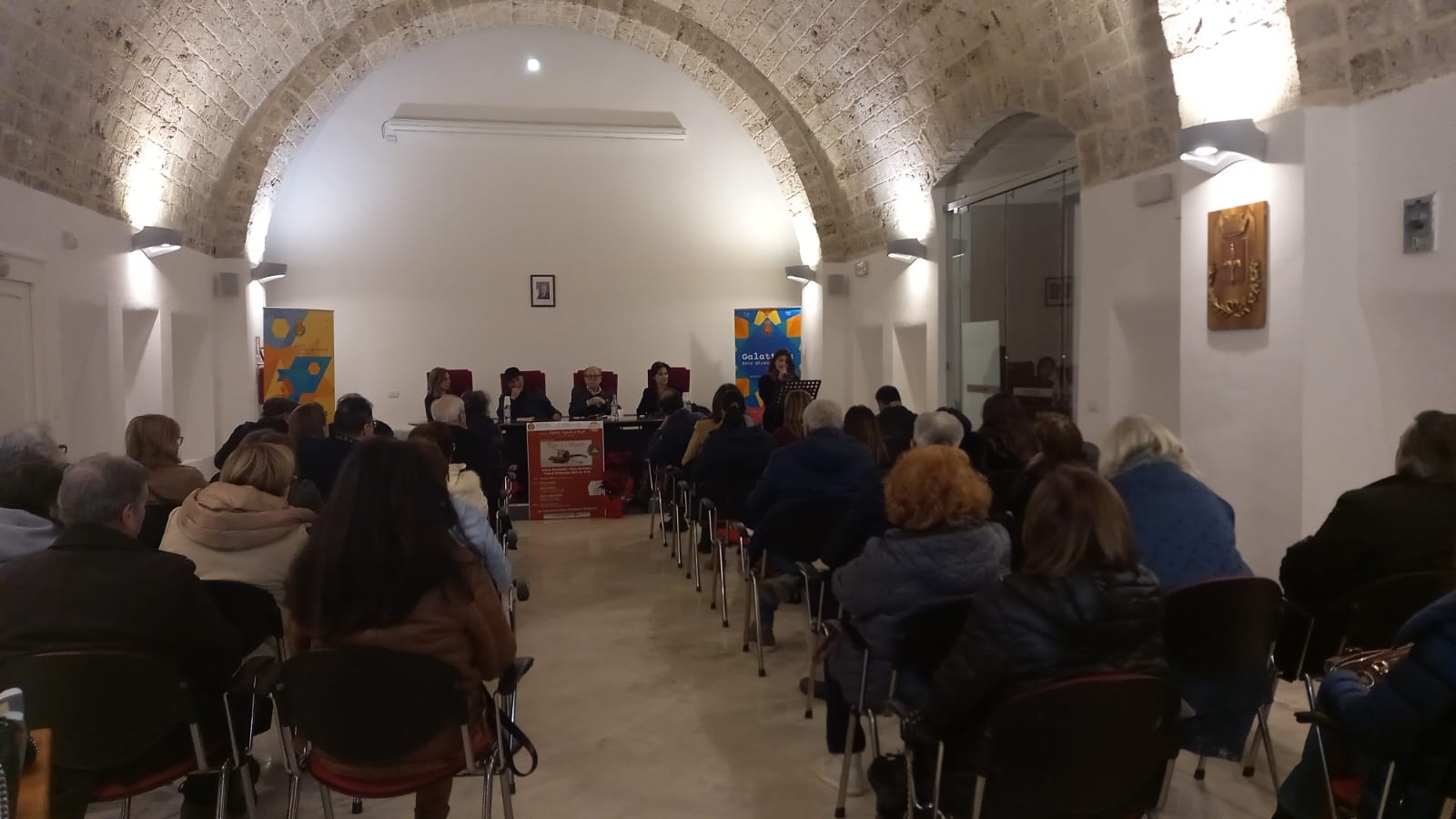 Platea di persone e tavolo centrale alla stanza. Dietro al tavolo sono sedute quattro persone e a fianco del tavolo, donna davanti a un leggio che legge qualcosa al microfono. Ai lati de tavolo ci sono due roll-up di Galattica.