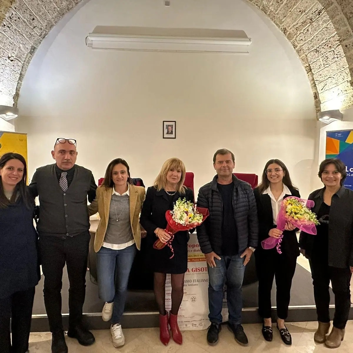 sette persone in piedi davanti a un tavolo sorridenti. Una ragazza e una signora tengono in mano un mazzo di fiori ciascuno