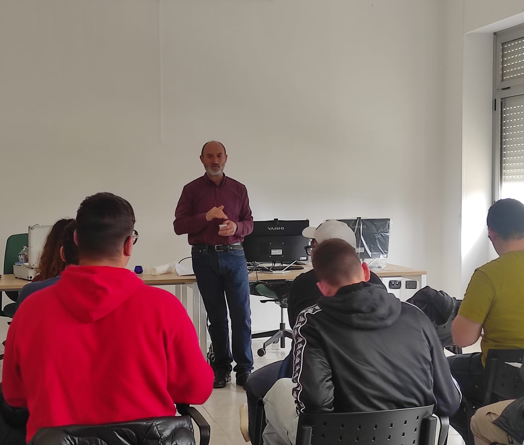 un gruppo di giovani stanno seduti di spalle, ascoltando un uomo in piedi di mezza età. l'umo sta davanti a un tavolo ed ha in mano un bicchiere piccolo.