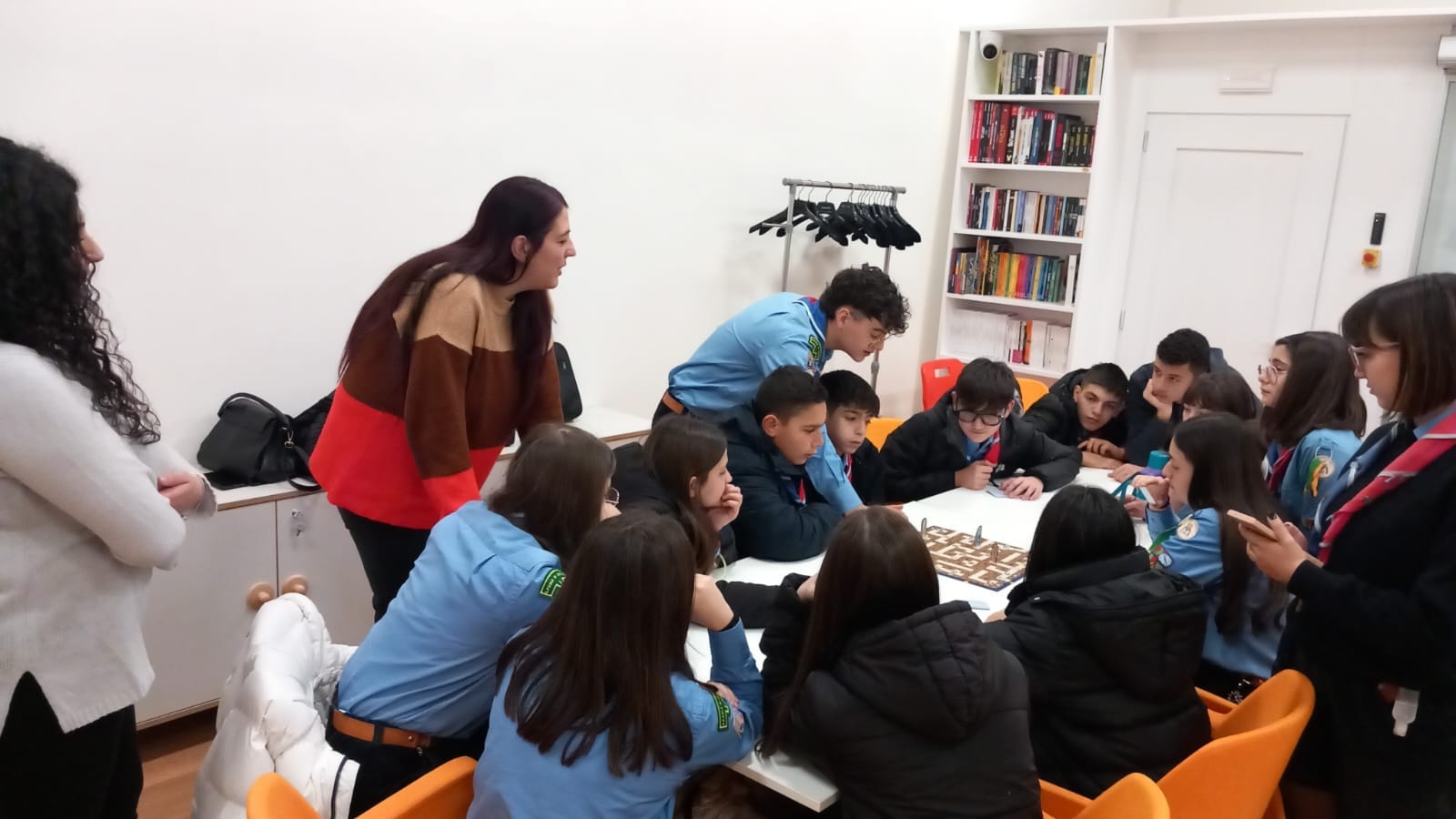 Alcuni agazzi seduti su sedie arancioni, altri ragazzi in movimento con uniforme scout, due ragazze in piedi, sullo sfondo muro bianco e parete con libri