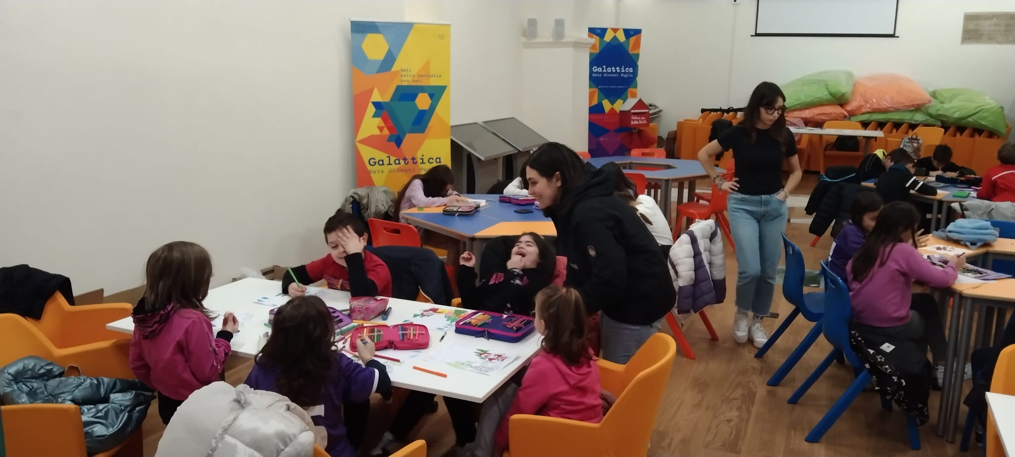 Bambini di spalle, con ragazza rivolta a parlare verso di loro. Parete bianca con roll-up arancione e blu di Galattica