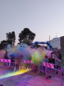 in mezzo a una strada ci sono delle transenne con dietro dei bambini che si intravedono in una nube di colori. Dietro di loro si intravede un arco blu di gomma con scritto AICS