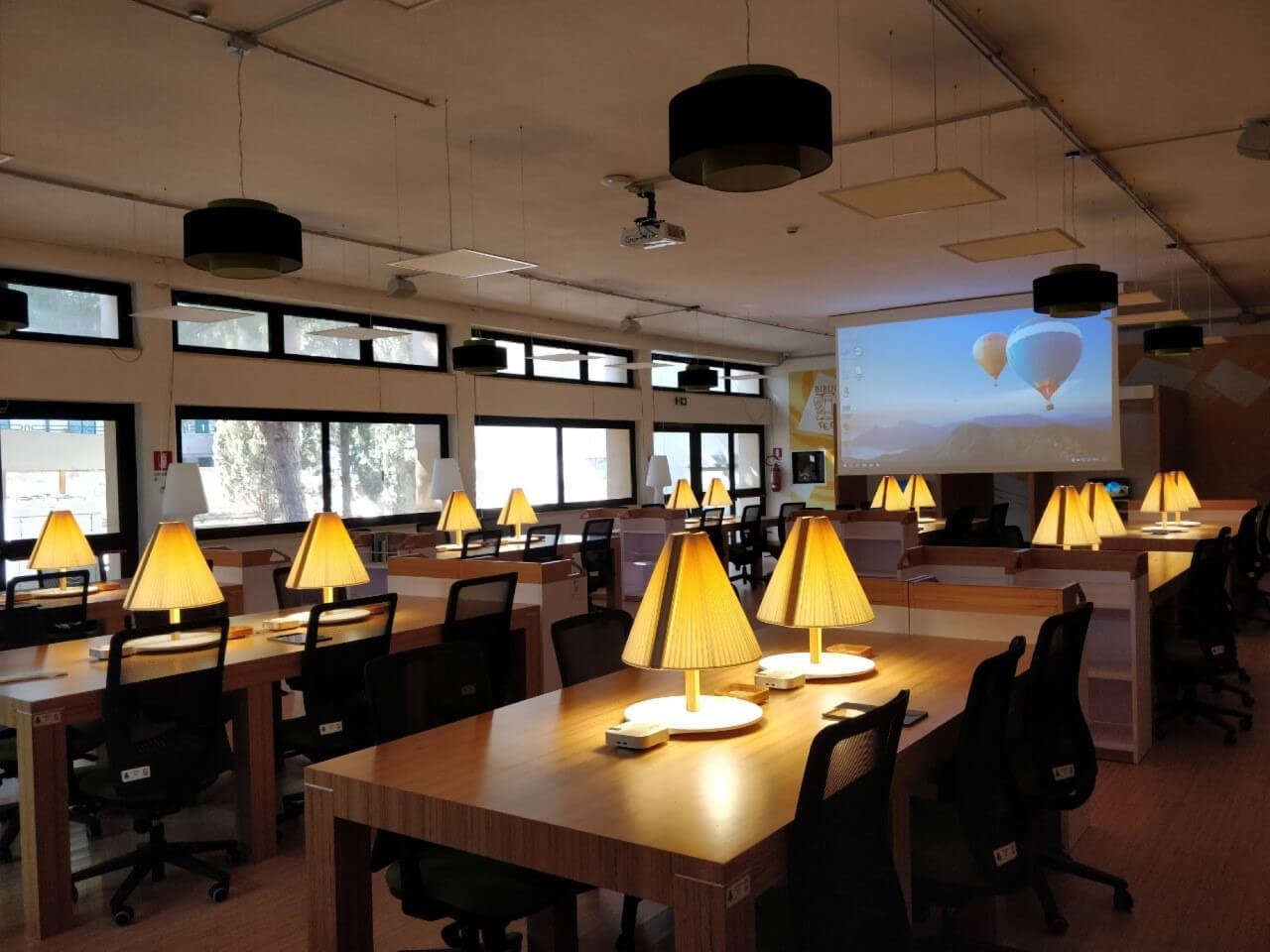 Sala lettura di una biblioteca con numerosi tavoli attrezzati e un grande schermo da proiezione