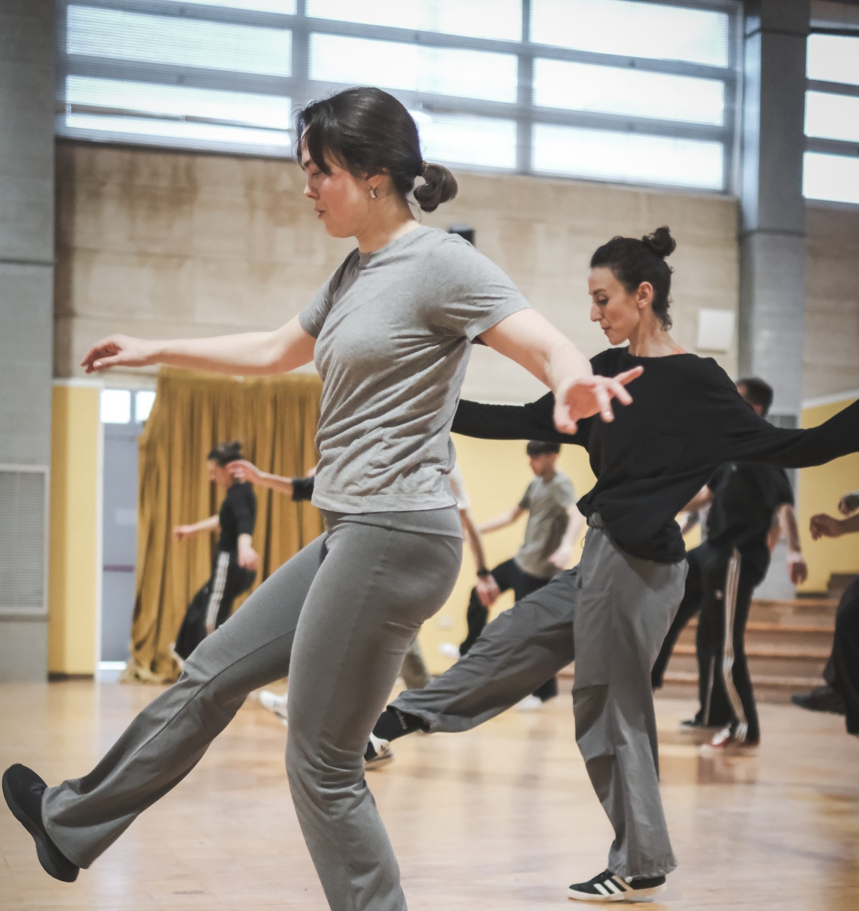 In primo piano due danzatrici alle prese con un passo di danza: una gamba su e una giù, bracia aperte per mantenere l'equilibrio. Dietro di loro, più sfocati, altri danzatori fanno la stessa cosa.