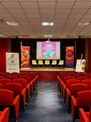 Teatro della sede del nodo di San Pancrazio Salentino Teatro con ai lati file di poltrone rosse, pavimento nero al centro e il palco di fronte in fondo con cinque sedie gialle e roll up di Galattica, locandina proiettata su sfondo bianco.