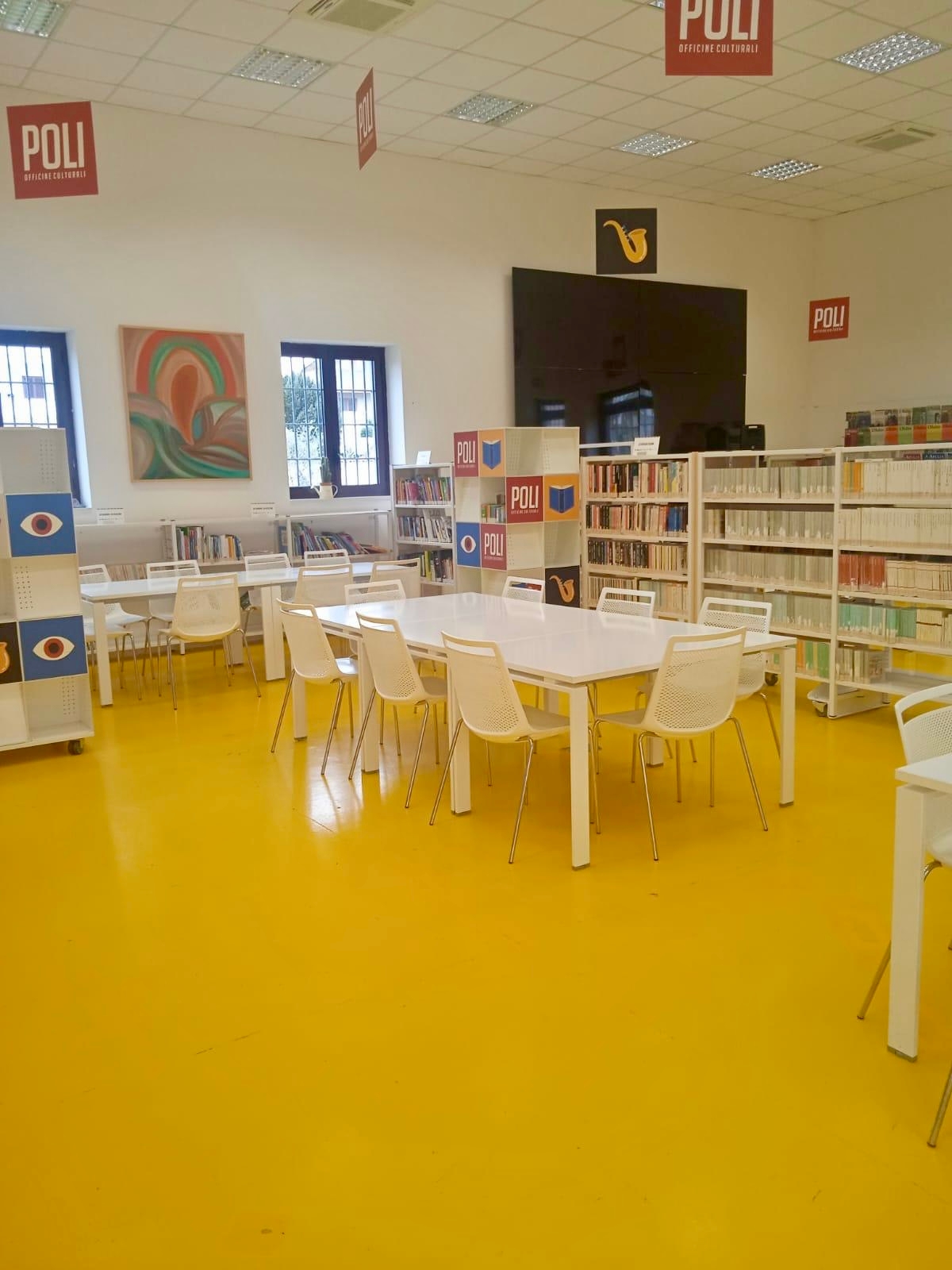 Pavimento giallo, sedie bianche attorno a un tavolo bianco centrale e un altro uguale sulla sinistra, scaffali pieni di libri, sullo sfondo un quadro colorato tra due finestre e un grande schermo nero a destra.