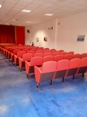 Teatro della sede del nodo di San Pancrazio Salentino Teatro con file di poltrone rosse e il palco con sipario rosso sullo sfondo.