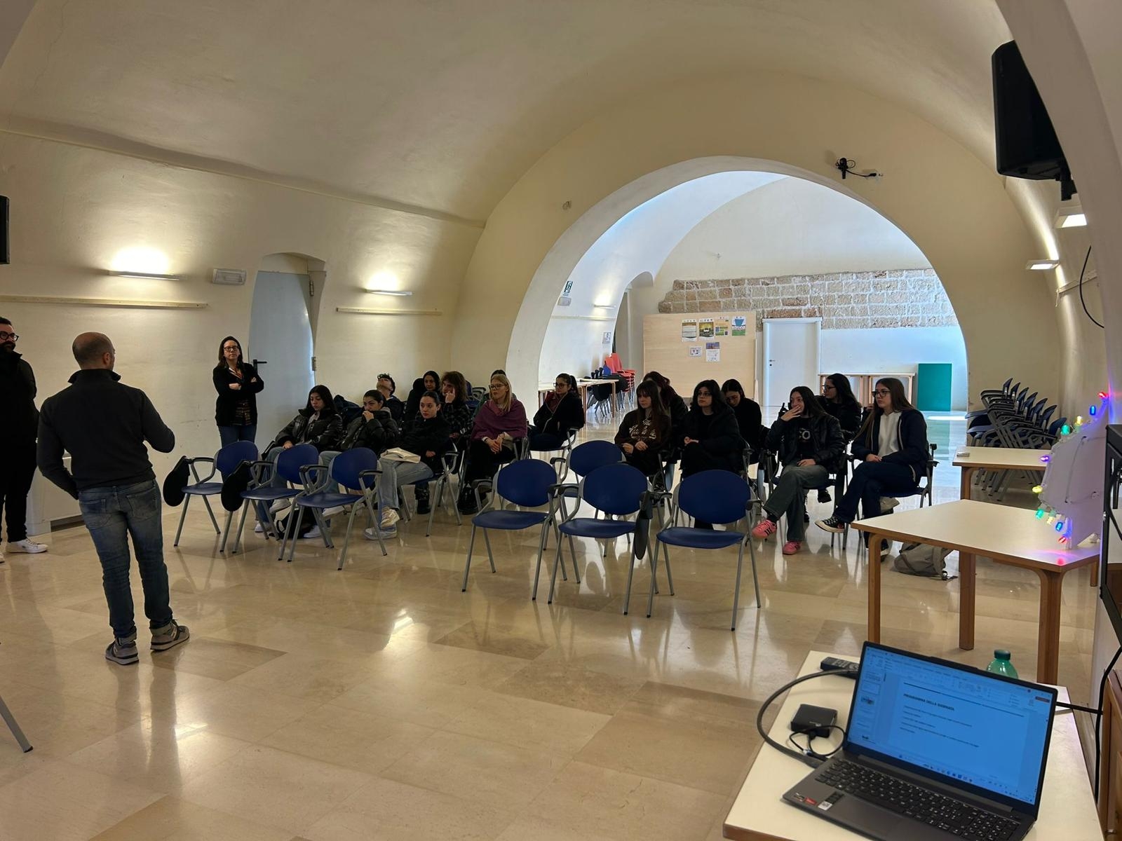 gruppo di giovani in una stanza ad archi che stanno ascotando un uomo in piedi che sta parlando di spalle. all'angolo destra dell'immagine c'è un pc aperto e dietro il computer una luminaria. Dall'altro lato della stanza a sinistra una donna e un uomo in piedi.