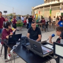 In primo piano un tavolo con sopra due pc e un mixer. Dietro al tavolo c'è un ragazzo seduto, girato di lato con gli occhiali che sta parlando al microfono che tiene in mano. Accanto a lui sulla sinistra un signore che sta guardando il ragazzo. Sul lato corto de tavolo, sta seduto un altro ragazzo che indossa delle cuffie audio, ha gli occhiali e tiene anche lui un microfono. Dietro di loro vari ragazzi alcuni seduti a dei tavoli, alcuni in piedi vicino ad una struttura sullo sfondo fatta da delle pareti a sbarre e colonne colorate.