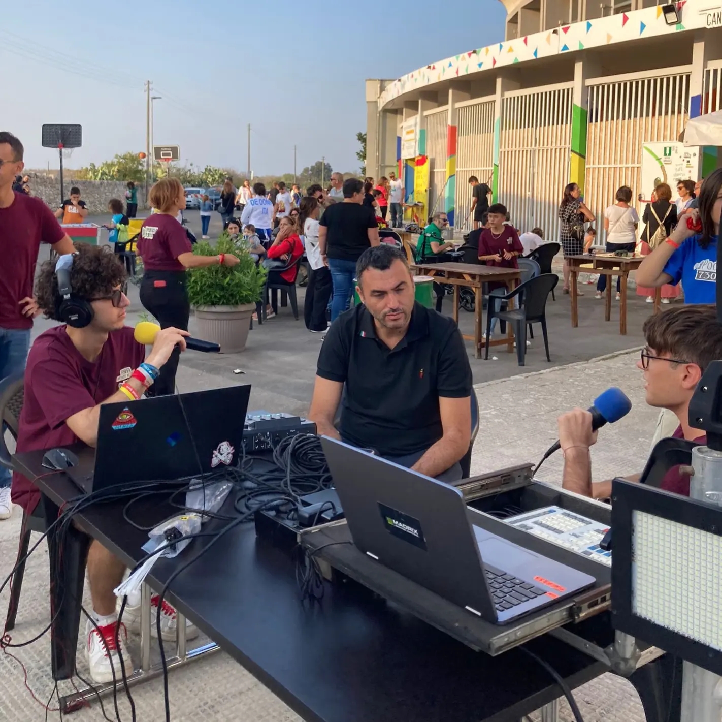 In primo piano un tavolo con sopra due pc e un mixer. Dietro al tavolo c'è un ragazzo seduto, girato di lato con gli occhiali che sta parlando al microfono che tiene in mano. Accanto a lui sulla sinistra un signore che sta guardando il ragazzo. Sul lato corto de tavolo, sta seduto un altro ragazzo che indossa delle cuffie audio, ha gli occhiali e tiene anche lui un microfono. Dietro di loro vari ragazzi alcuni seduti a dei tavoli, alcuni in piedi vicino ad una struttura sullo sfondo fatta da delle pareti a sbarre e colonne colorate.