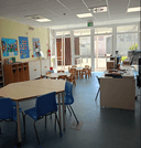 interno vuoto 2 Presenza di un tavolo posto al centro dell'immagine e adiacenti ci sono altri due tavoli più piccoli per bambini ogni tavolo con sei sedioline. E' presente un mobiletto sul lato sinistro e al di sopra sono presenti dei cartelloni decorati dai bambini della scuola materna del nodo. Sul lato destro invece troviamo la postazione dell'operatore. In fondo alla sala è presente una vetrata che affaccia all'esterno dello spazio dove si intravede la book house e il giardino.