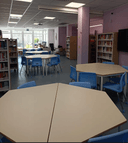 interno biblioteca Panoramica della biblioteca : presenza di tavoli a formare un esagono nel quale è posta una sedia per lato per un totale di sei sedie. Presenza di scaffali sia sul lato destro che sinistro e in mezzo allo spazio fra i tavoli è presente un pilastro. In fondo alla stanza è posta una postazione sede dell'operatore e ampia vetrata.