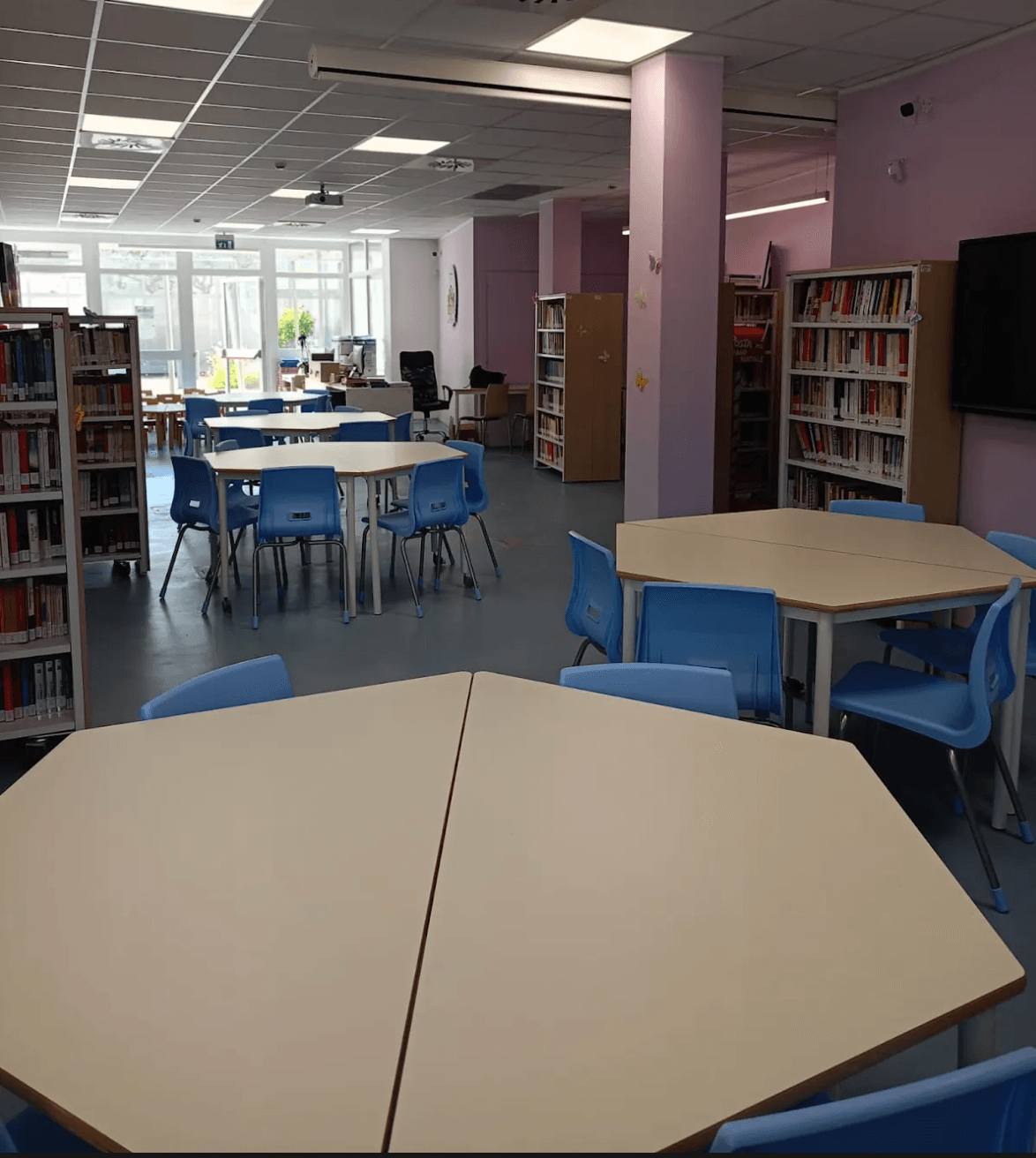 Panoramica della biblioteca : presenza di tavoli a formare un esagono nel quale è posta una sedia per lato per un totale di sei sedie. Presenza di scaffali sia sul lato destro che sinistro e in mezzo allo spazio fra i tavoli è presente un pilastro. In fondo alla stanza è posta una postazione sede dell'operatore e ampia vetrata.