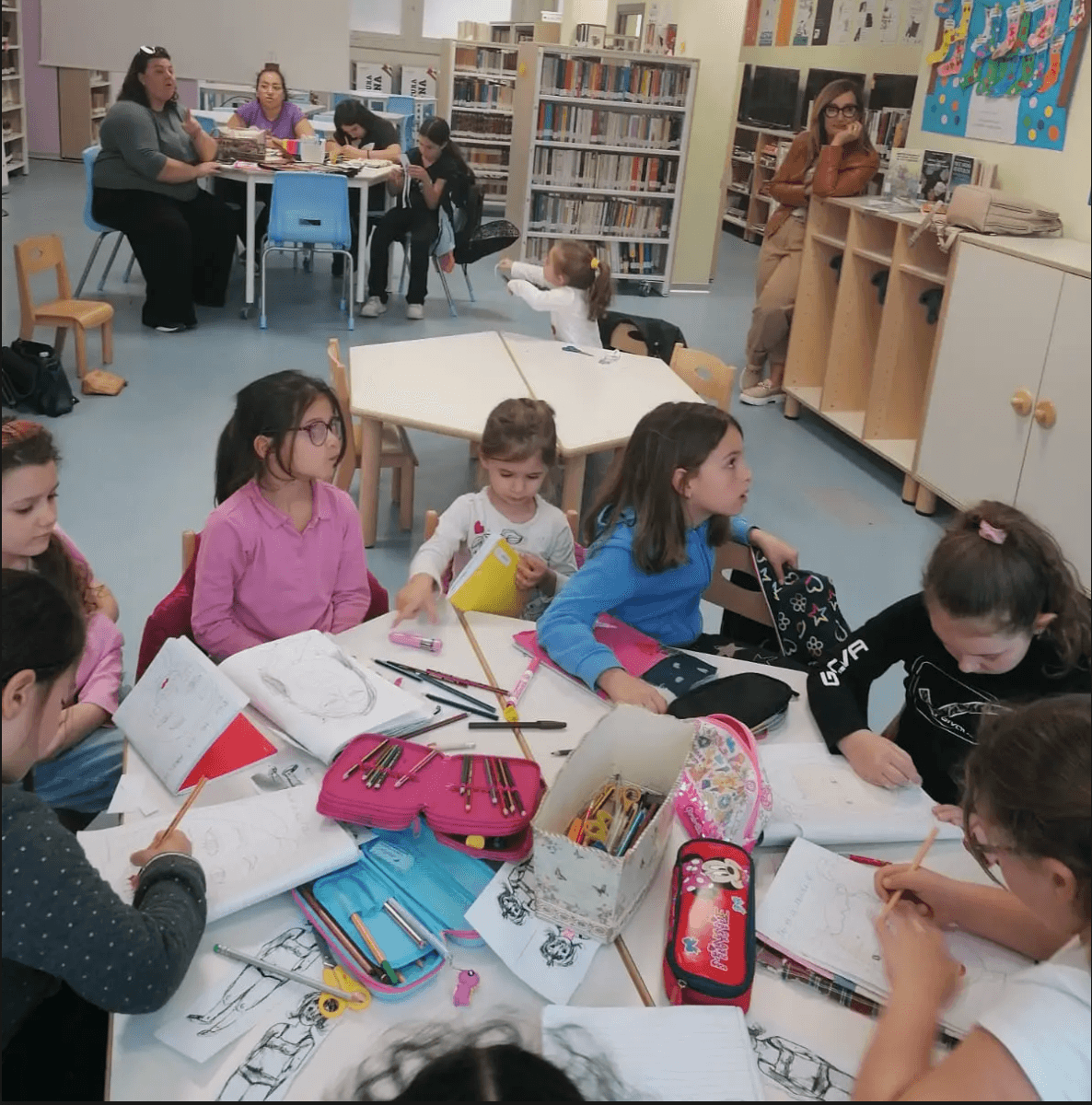 Nella foto sono presenti dei bambini seduti sulle sedioline mentre sono impegnati nell'attività di disegno. I bambini sono acompagnati dai genitori posti alle loro spalle sul tavolo più grande circondati dgli scaffali.