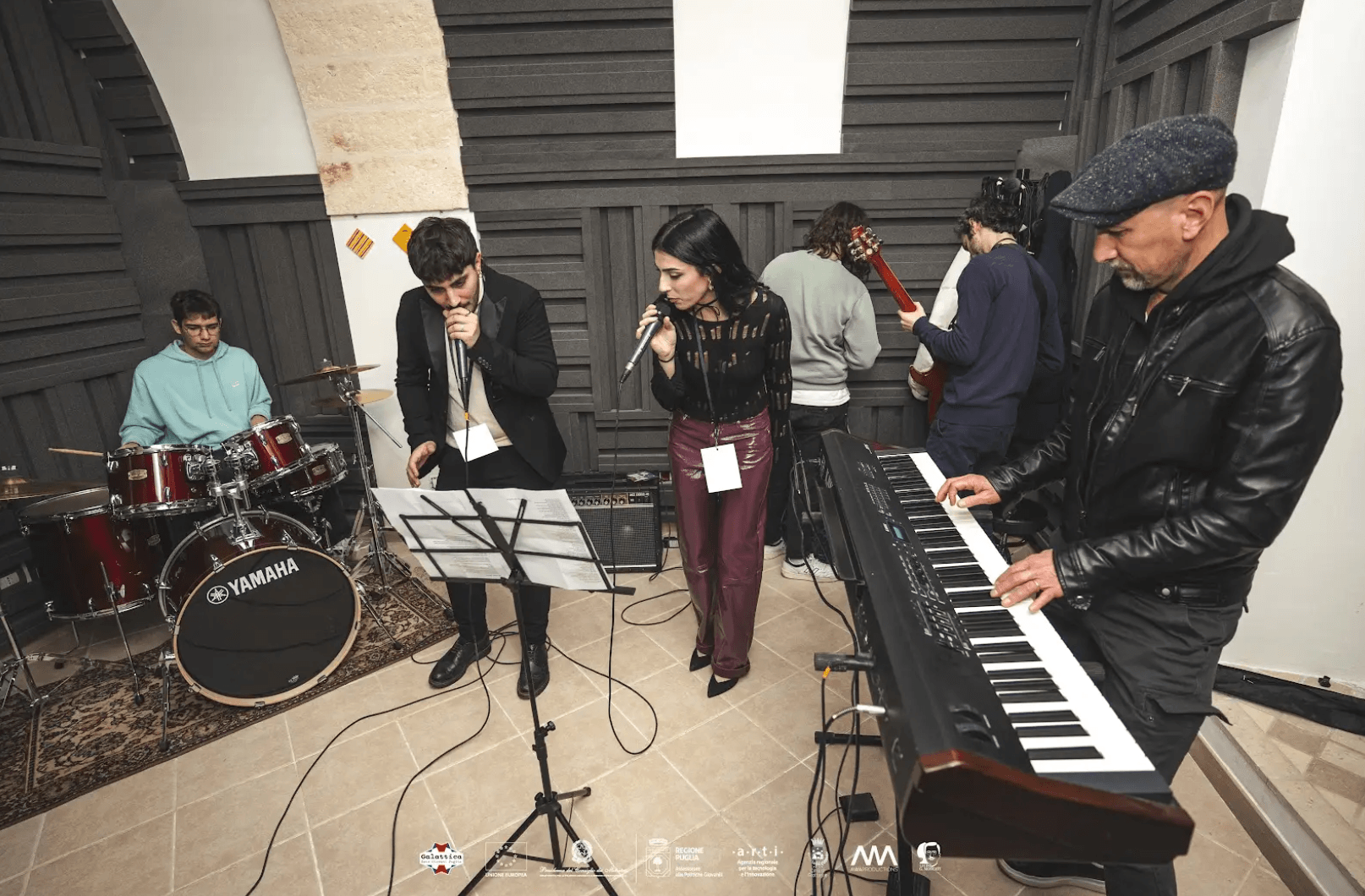 Sala Produzione Musicale in attività. Nella foto sono presenti sei persone in una stanza insonorizzata, impegnate in una sessione musicale. A sinistra c'è un batterista seduto dietro un set di batteria Yamaha rosso. Indossa una felpa verde acqua. Al centro ci sono due cantanti: un uomo con una giacca nera e pantaloni scuri, che tiene un microfono e legge da un leggio, e una donna con una maglia nera trasparente e pantaloni di pelle viola scuri, che canta in un altro microfono. Sulla destra, un uomo con un cappello piatto grigio e una giacca di pelle nera suona una tastiera elettronica. In secondo piano, due altri musicisti: uno, con una maglia verde e jeans, è rivolto verso il muro e sembra prepararsi a suonare una chitarra elettrica, mentre l'altro, con una maglia viola e jeans, è impegnato con un altro strumento a corda. Il pavimento è di piastrelle chiare e sulle pareti ci sono pannelli fonoassorbenti. In basso al centro dell'immagine, ci sono vari loghi e scritte, tra cui "Galattica".