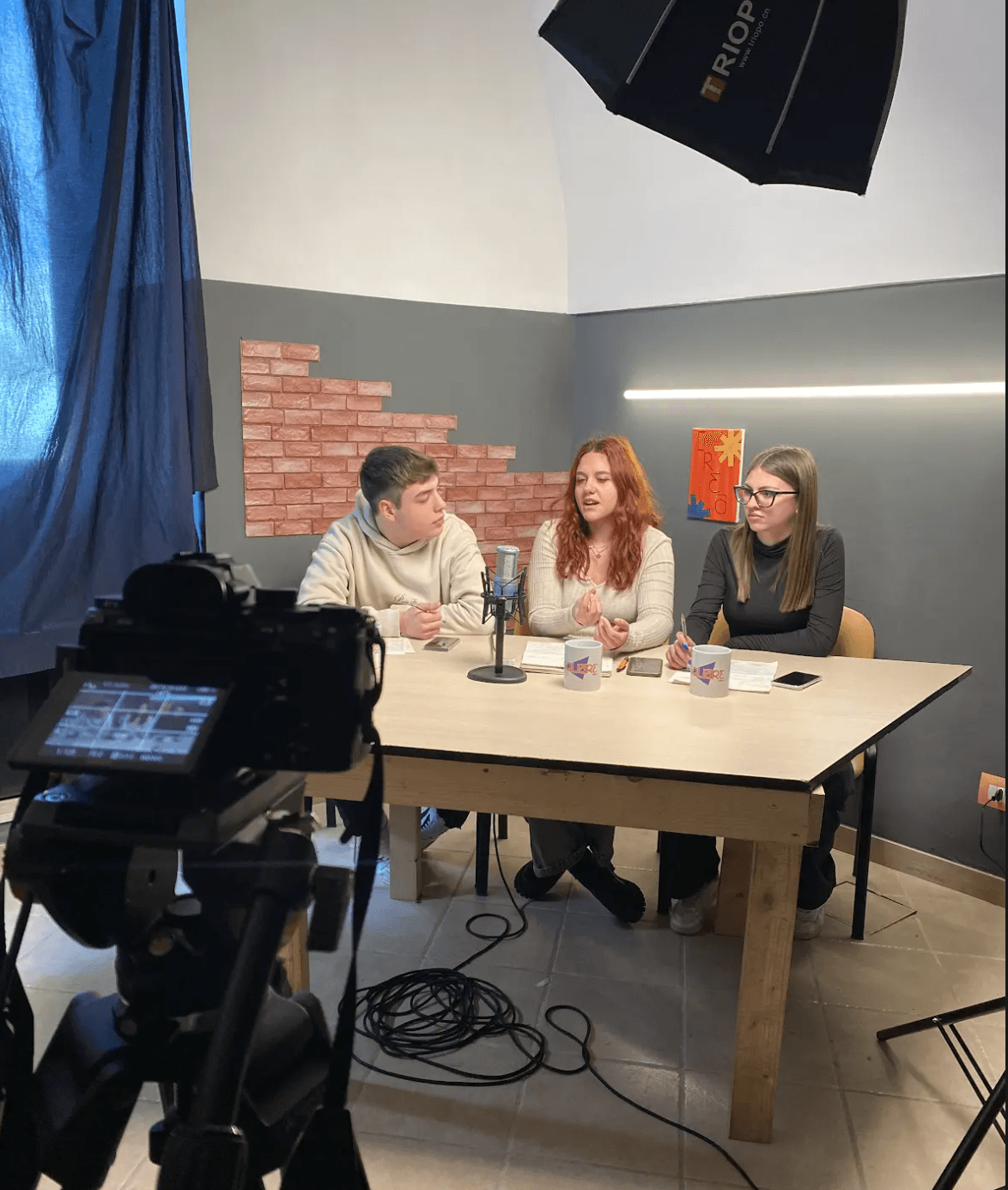Nella foto ci sono tre persone sedute a un tavolo di legno chiaro. Sul tavolo ci sono tazze bianche con la scritta "TheSquare", uno smartphone e dei fogli. Al centro del tavolo è posizionato un microfono. La persona a sinistra indossa una felpa chiara e guarda verso la persona al centro, che ha i capelli rossi e indossa un maglione chiaro. La persona a destra ha i capelli castani lunghi e porta gli occhiali, indossa un maglione scuro. Davanti al tavolo, in primo piano, c'è una videocamera montata su un treppiede, con un cavo che si estende sul pavimento piastrellato. Sopra il tavolo c'è un grande riflettore. La parete di fondo ha un pannello di finta muratura rossa e un quadro colorato con scritto "CREA!". La stanza è parzialmente illuminata dalla luce proveniente da una finestra coperta da una tenda scura sulla sinistra.