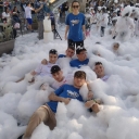 Alcuni ragazzi stesi per terra tra la schiuma, una ragazza in piedi con gli occhiali da sole. Dietro di loro tanti altri ragazzi con la schhiuma sulla testa. In piedi sulla destra si intavede una figura maschile con un microfono. Davanti a lui una contenitore di plastica.