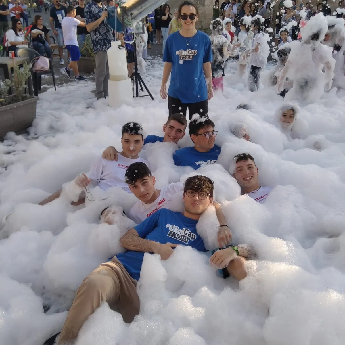 Alcuni ragazzi stesi per terra tra la schiuma, una ragazza in piedi con gli occhiali da sole. Dietro di loro tanti altri ragazzi con la schhiuma sulla testa. In piedi sulla destra si intavede una figura maschile con un microfono. Davanti a lui una contenitore di plastica.